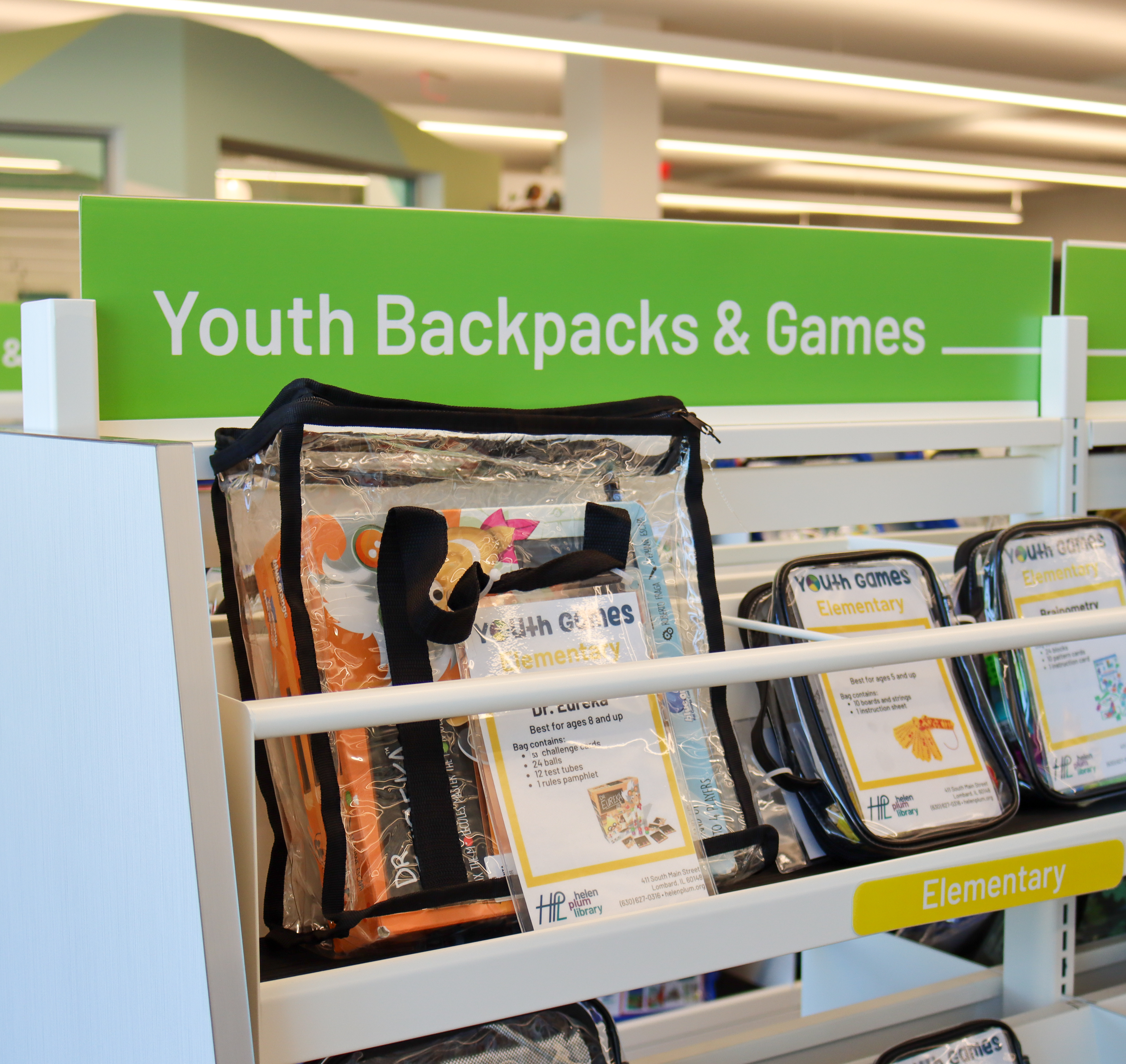 Shelf in Youth Services with a green sign that says Youth Backpacks & Games. Under the sign are 3 backpacks with various youth games inside.
