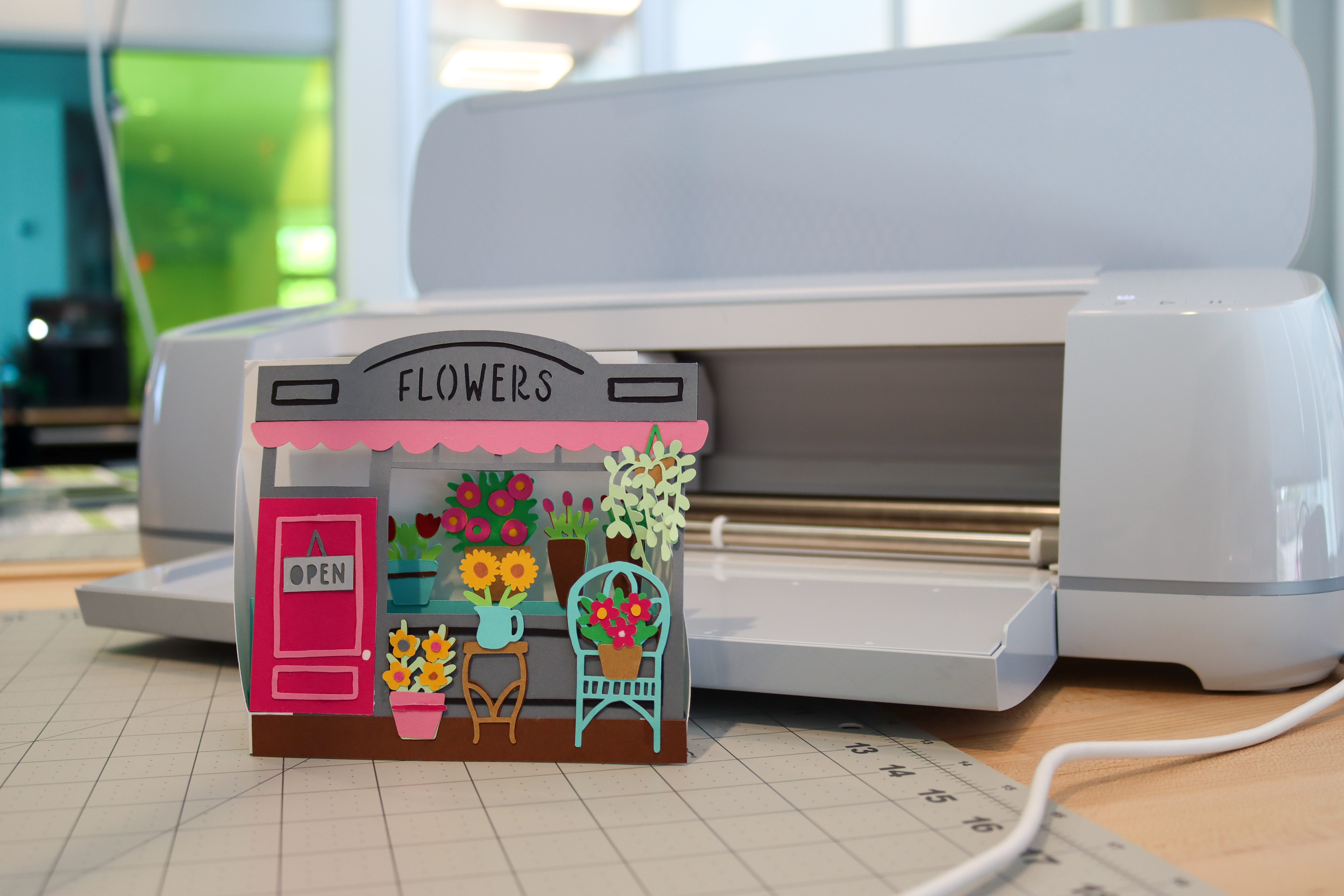 A 3D greeting card standing on a table in front of a Cricut machine. The card is a flower shop the a bright pink door and pots of flowers in the window and outside the shop.