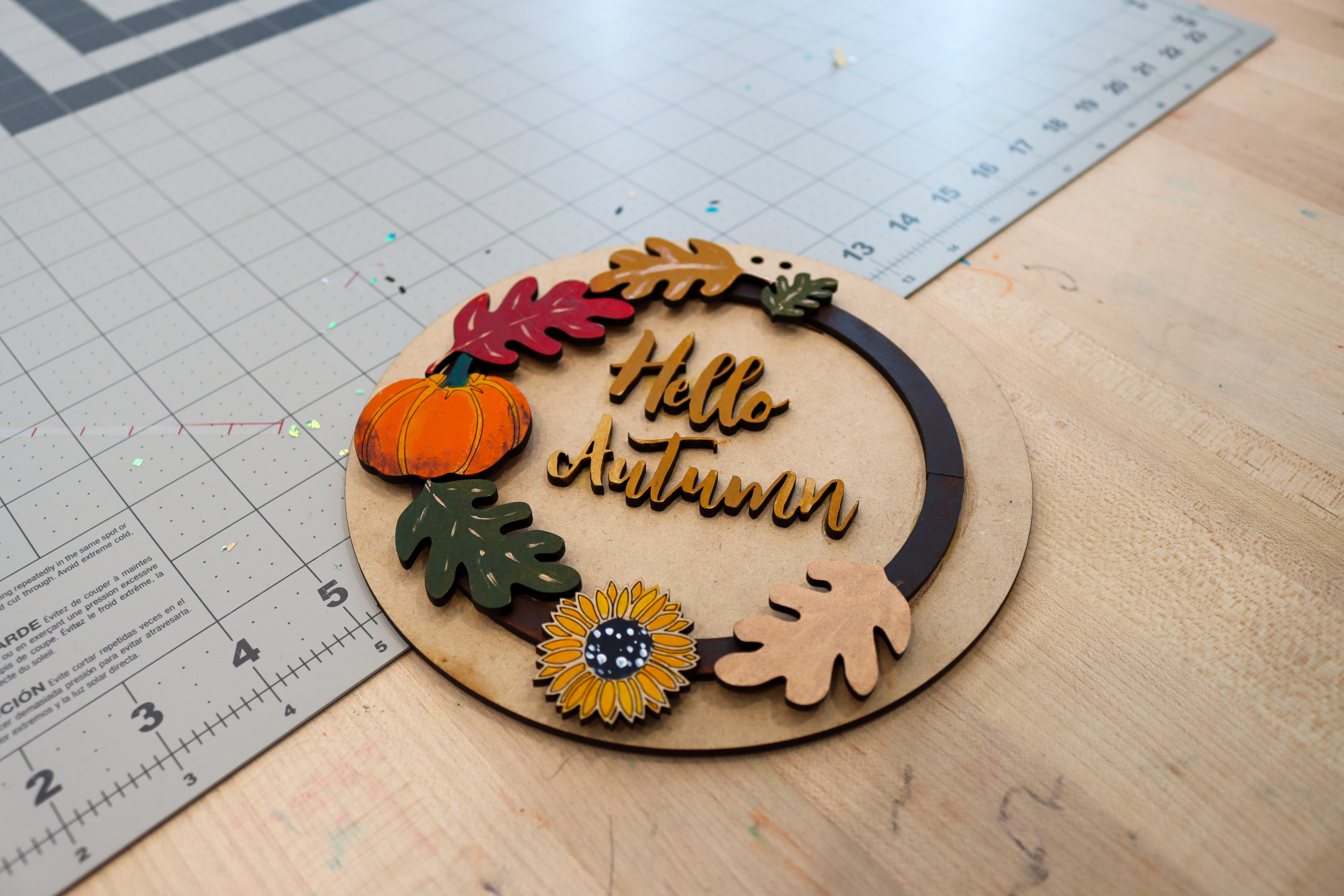 Circular sign made of wood that says hello, autumn in a wooden script. Around the words is a decorative wooden wreath with leaves and a pumpkin. 